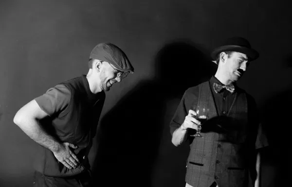 Two men in flat caps, one lighting a cigarette, lit hard against a dark background