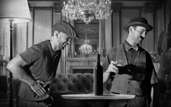The same two men in a hotel lounge under a chandelier, in black and white