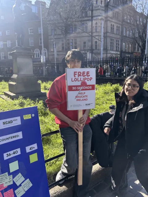 A team member holding a “Free lollipop, 30 seconds” sign next to the research board in a park