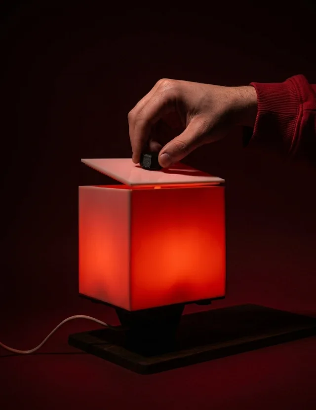 A hand lifting the lid of NOX, the cube glowing red on its wooden base
