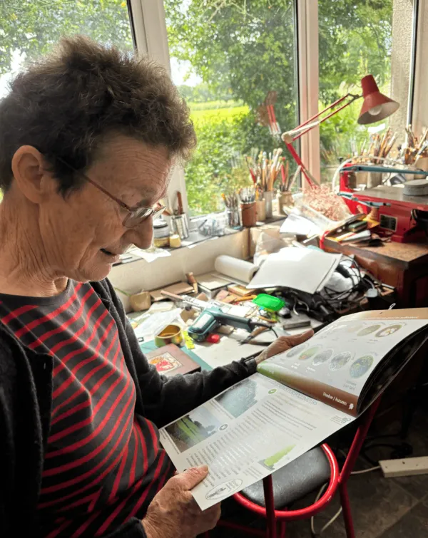 A maker reading printed papers at her desk, her studio shelves and tools around her